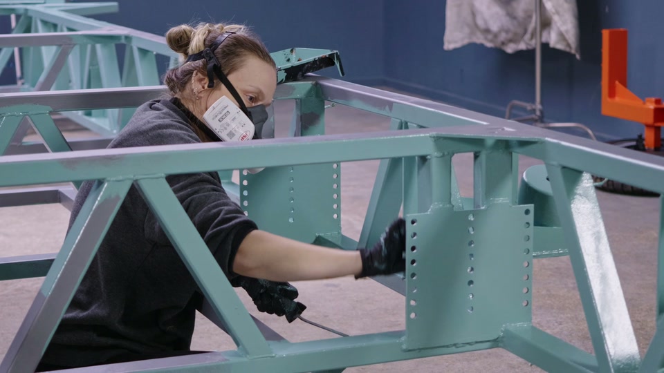 Hayley in a respirator reaching a long fine brush inside a box section of the tiny house trailer chassis