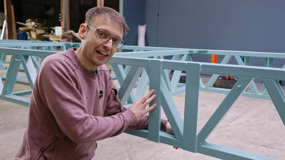 Pete resting a hand on the teal tiny house trailer chassis rail where the hydraulic leg brackets used to be
